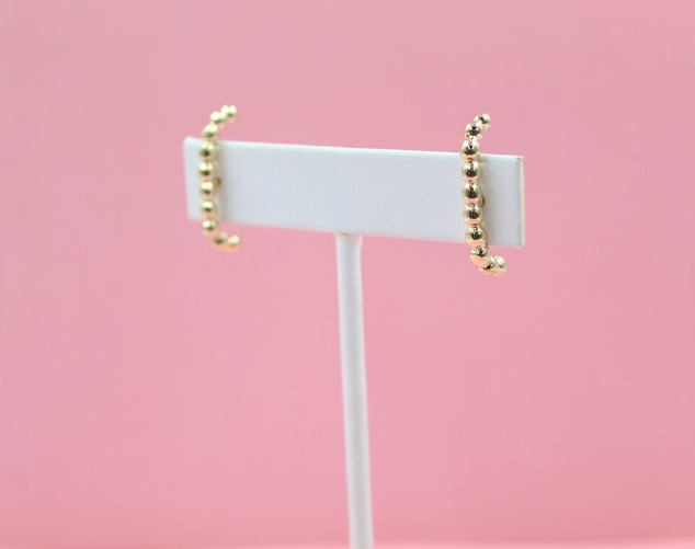 A pair of beaded gold crawler earrings rests on a white display stand against a soft pink background.