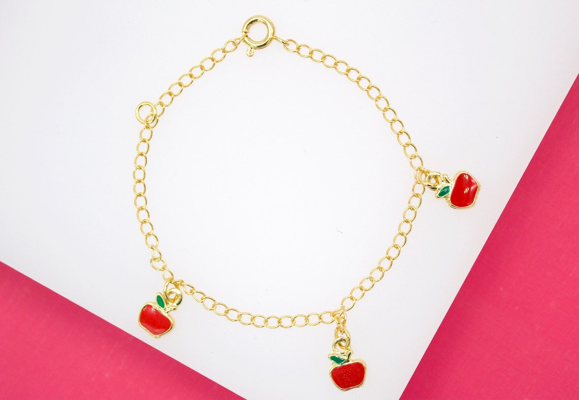 A delicate gold chain bracelet features three charming red enamel apple charms dangling against a white and pink backdrop.