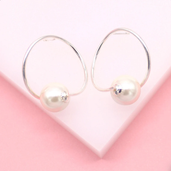 A pair of elegant silver hoop earrings featuring dangling white pearls rests against a soft pink backdrop.