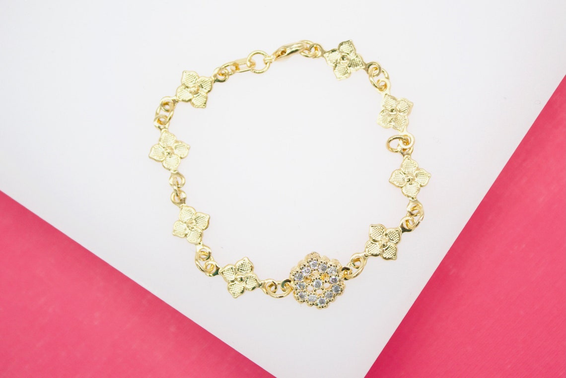 A delicate gold chain bracelet features clover-shaped links and a sparkling diamond-encrusted flower charm resting on a white and pink surface.