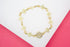 A delicate gold chain bracelet features clover-shaped links and a sparkling diamond-encrusted flower charm resting on a white and pink surface.