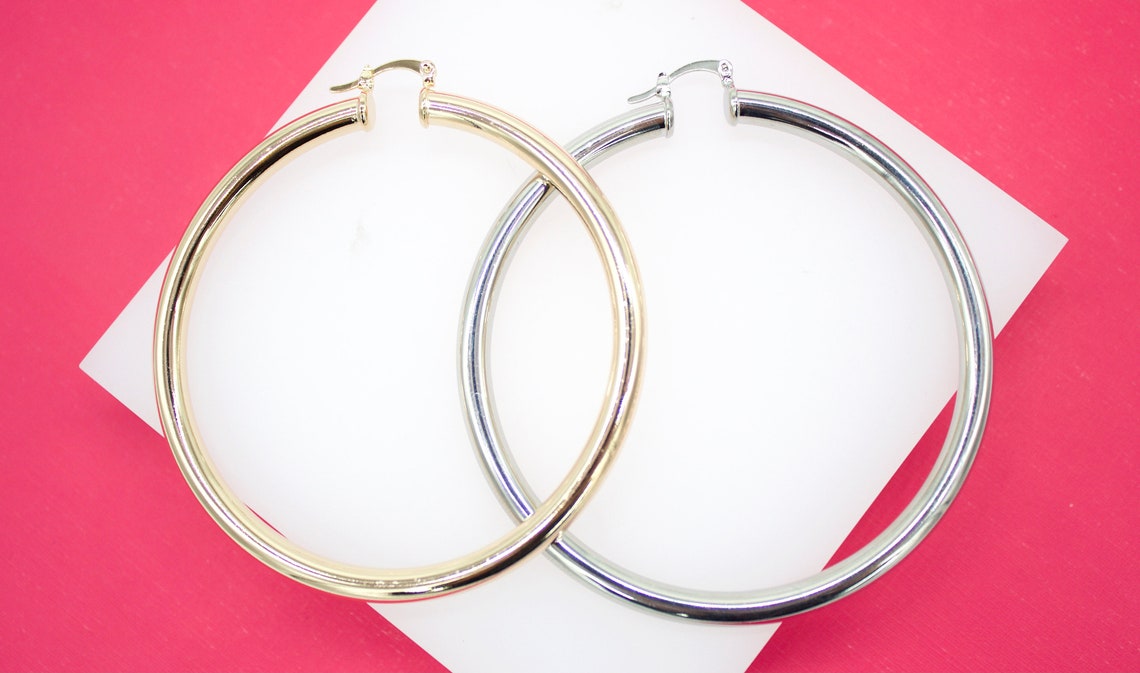 A pair of large gold and silver hoop earrings rests on a white display against a vibrant pink background.