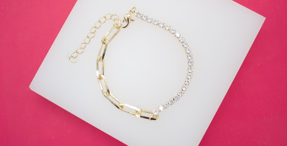 A gold half-paperclip and half-diamond tennis bracelet rests on a white display against a vibrant pink background.