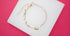 A gold half-paperclip and half-diamond tennis bracelet rests on a white display against a vibrant pink background.