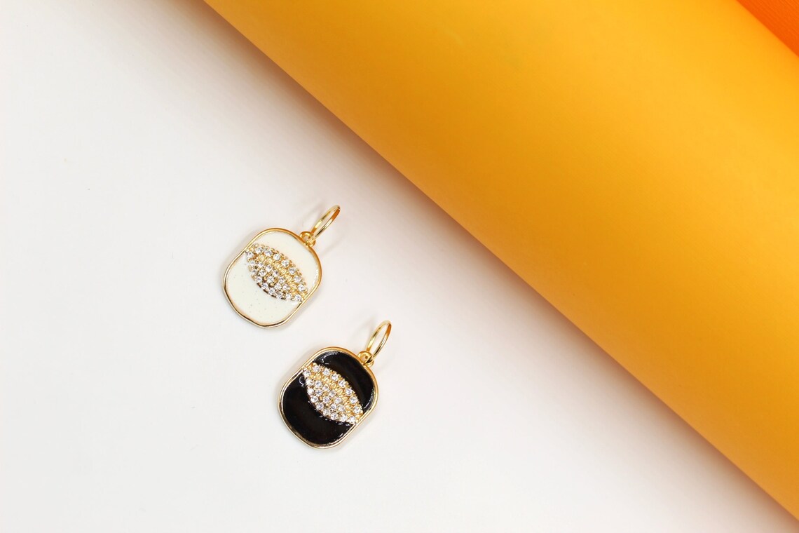 Two gold-plated enamel pendants featuring pave crystal evil eye motifs rest on a white surface against an orange and yellow background.