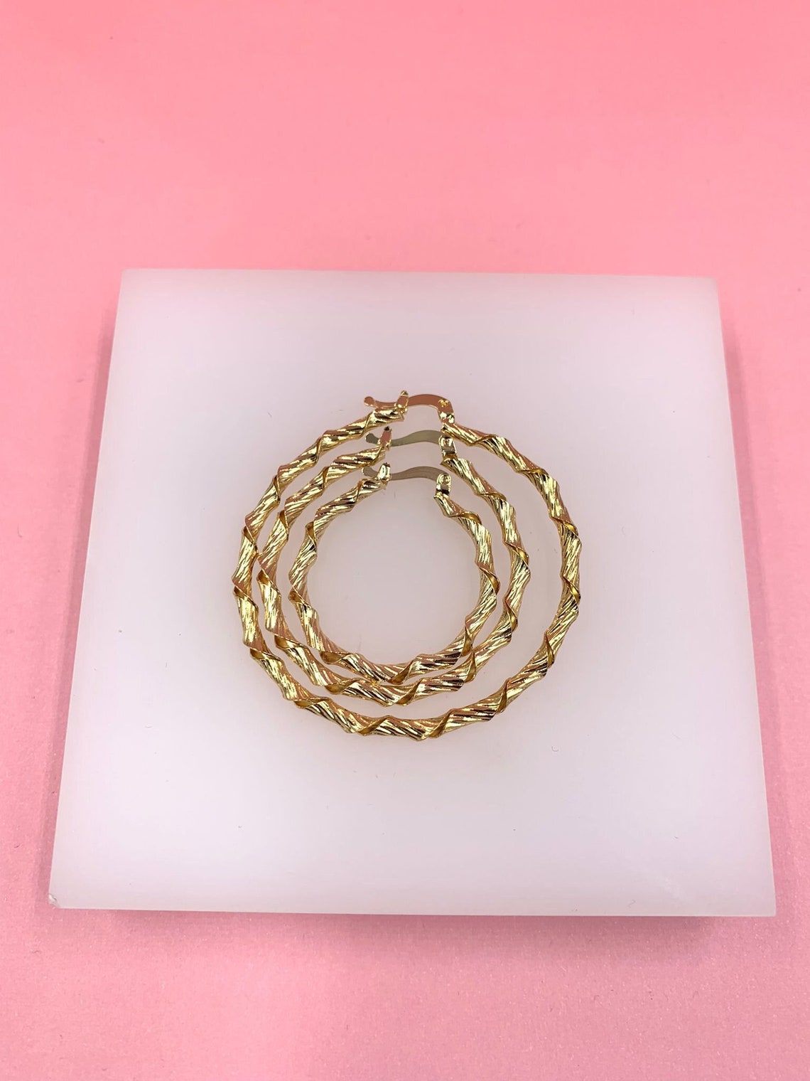 Three textured gold hoop earrings rest nested inside each other on a white square display tile against a pink background.