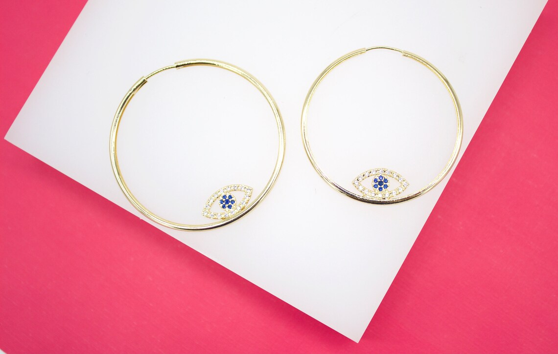 A pair of gold evil eye hoop earrings rests on a white and pink display surface.