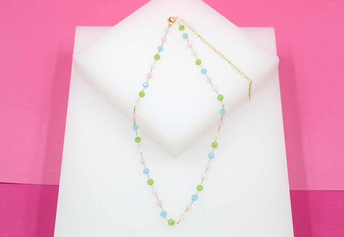 A delicate gold beaded necklace featuring colorful pastel green, pink, blue, and white stones rests on a white display block against a pink background.