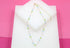 A delicate gold beaded necklace featuring colorful pastel green, pink, blue, and white stones rests on a white display block against a pink background.
