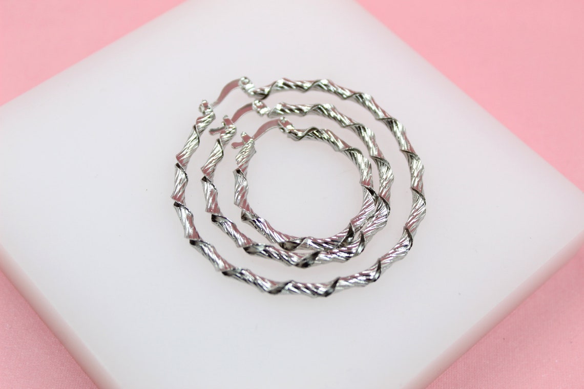 Three nested silver hoop earrings with a twisted, textured design rest on a white display surface against a pink background.