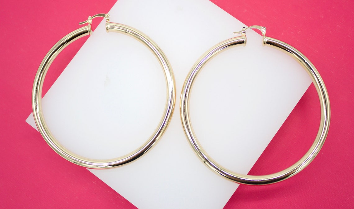 A pair of polished gold hoop earrings rests against a white display block on a vibrant pink background.