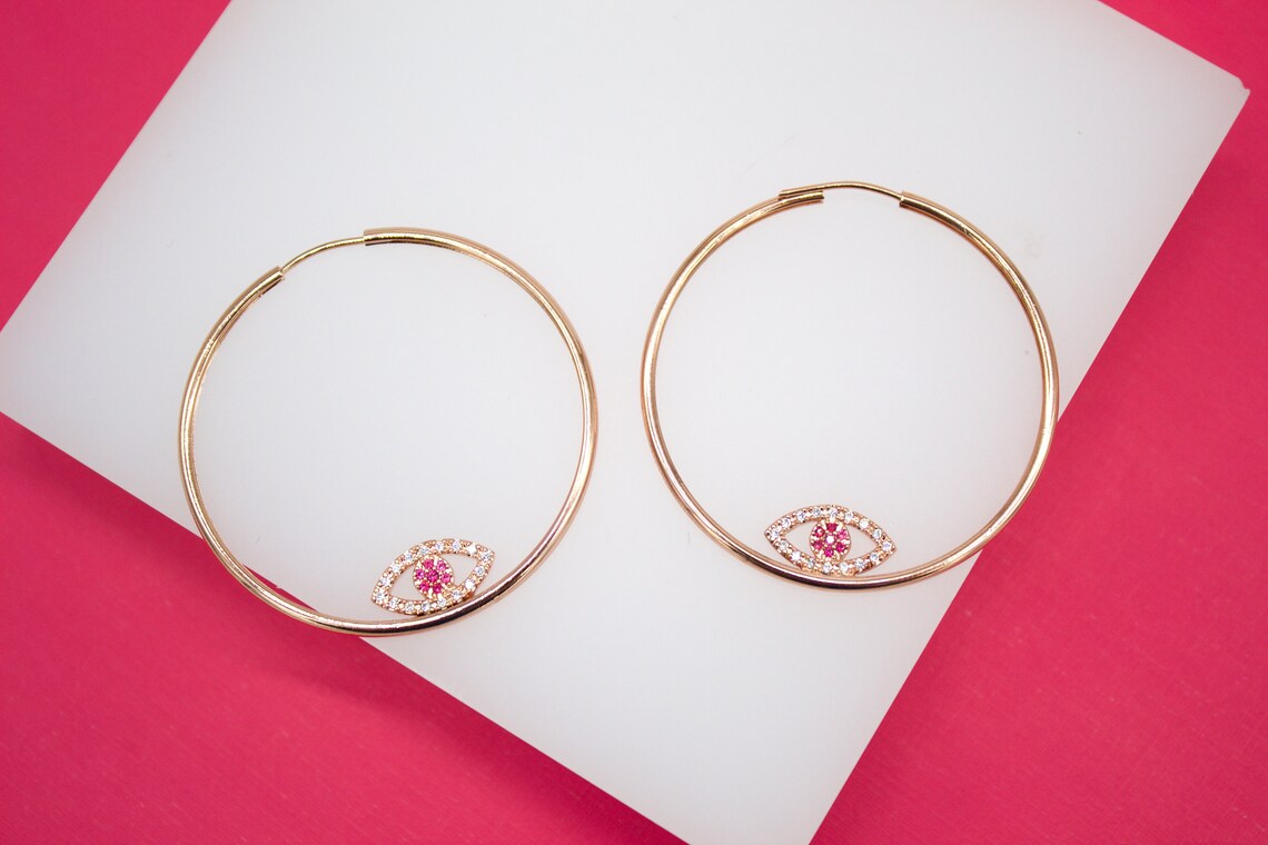 A pair of gold hoop earrings featuring evil eye charms with pink and white cubic zirconia stones rest on a white display against a vibrant pink background.