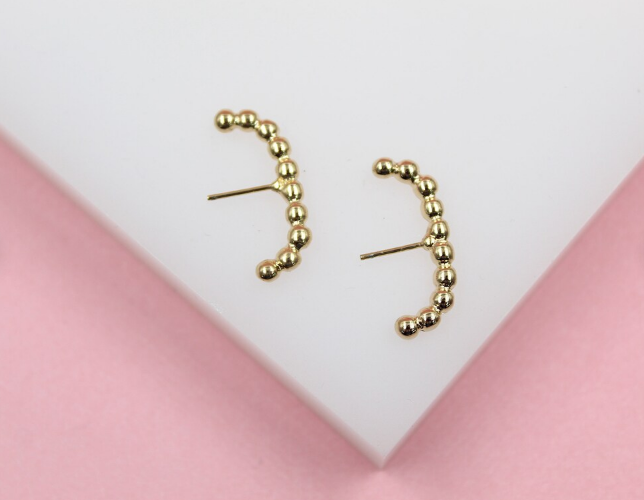 A pair of curved gold bead climber earrings rests on a white display surface against a soft pink background.
