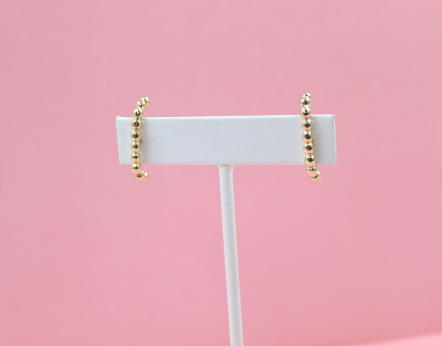 A pair of gold beaded climber earrings rests on a white display card against a soft pink background.