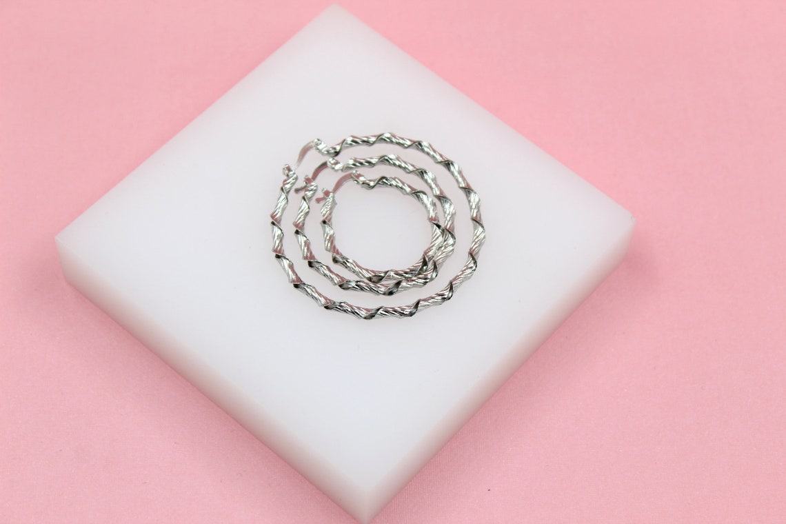 Three nested silver twisted hoop earrings rest on a white display block against a soft pink background.