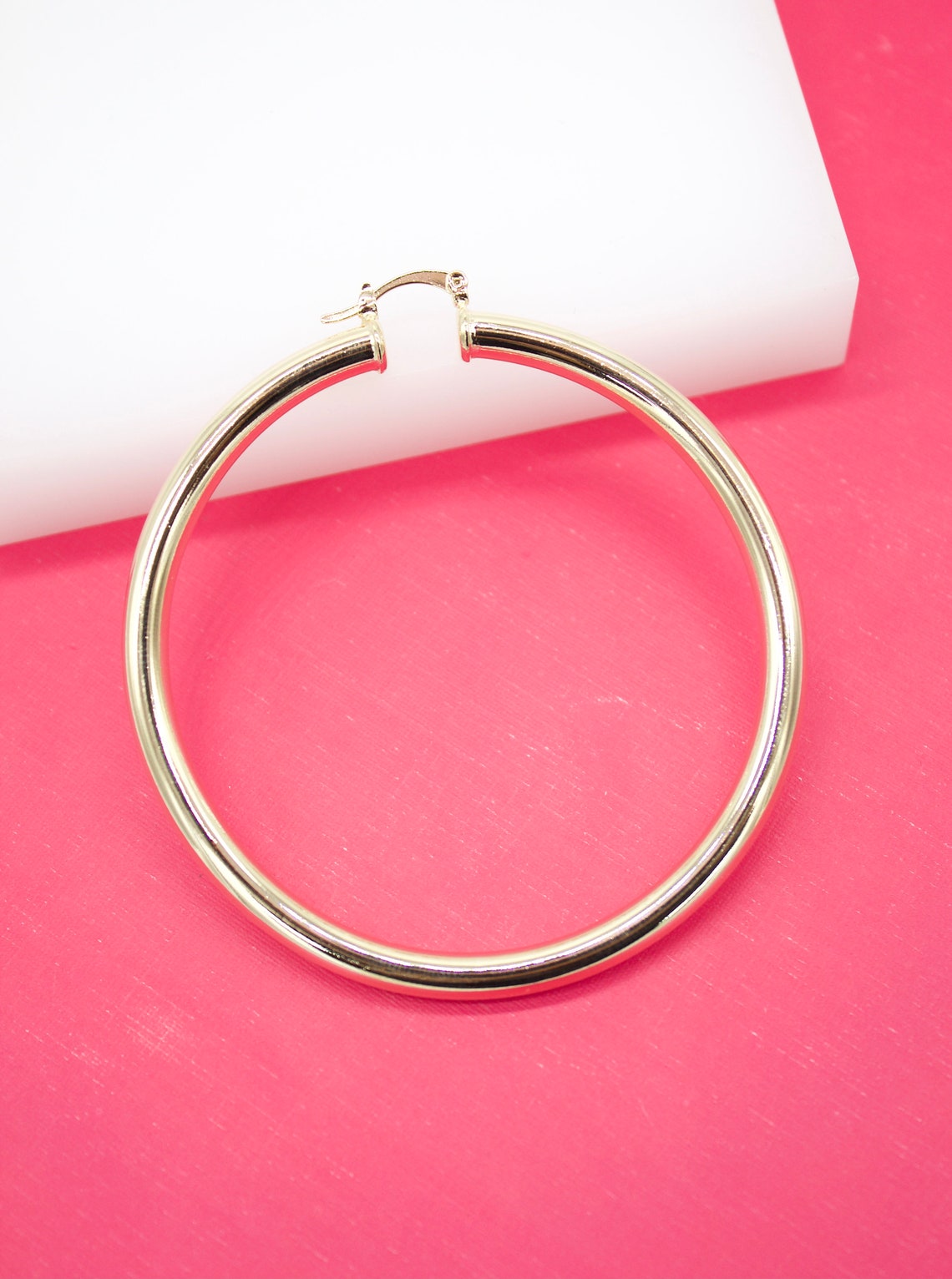 A polished gold hoop earring rests on a bright pink surface against a white backdrop.