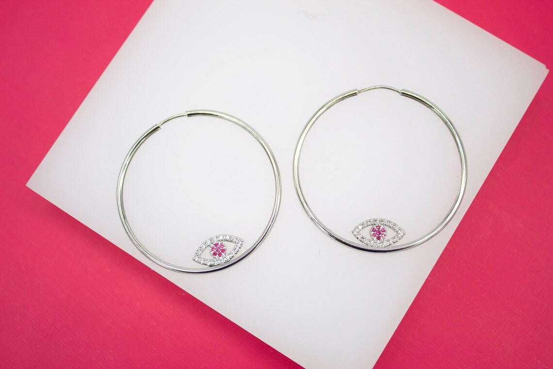 Two silver hoop earrings featuring sparkling crystal and pink gemstone evil eye charms rest on a white surface against a bright pink background.