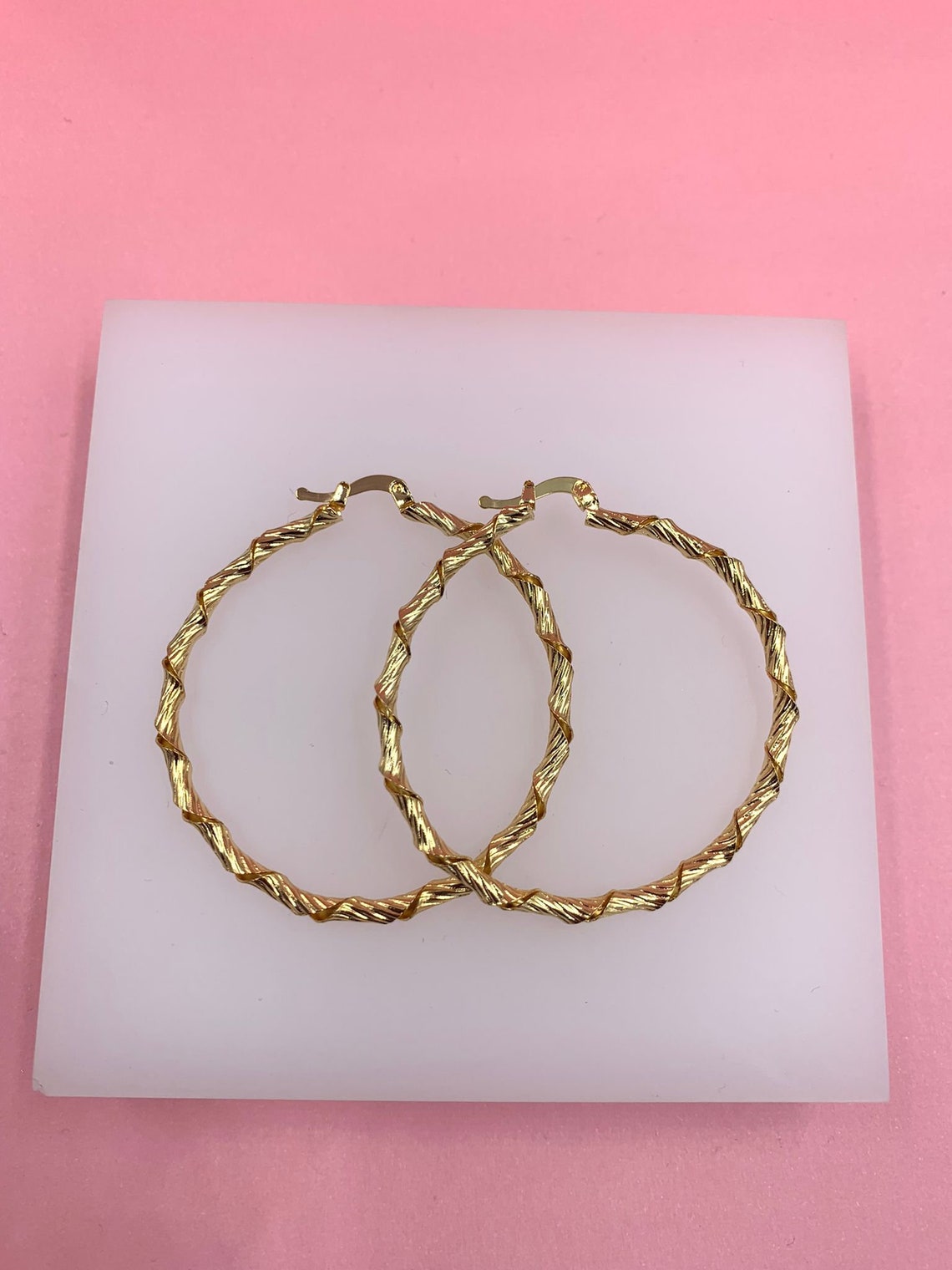 Two large, twisted gold hoop earrings rest on a white display block against a pink background.