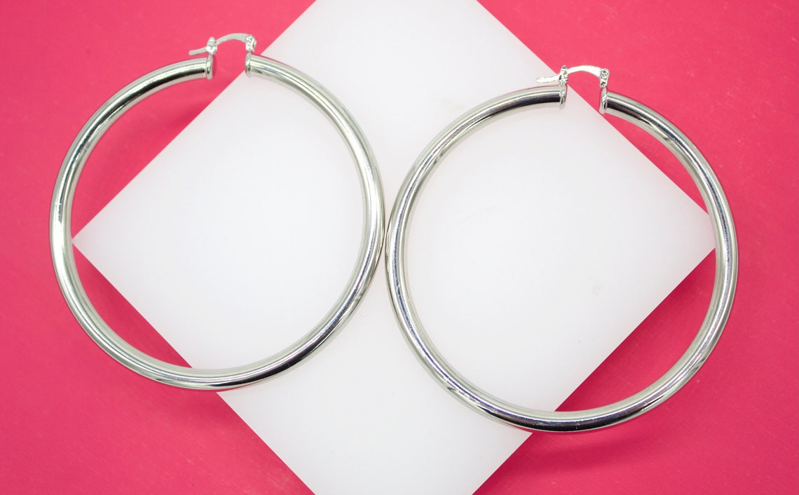 A pair of sleek silver hoop earrings rests on a white display against a vibrant pink background.