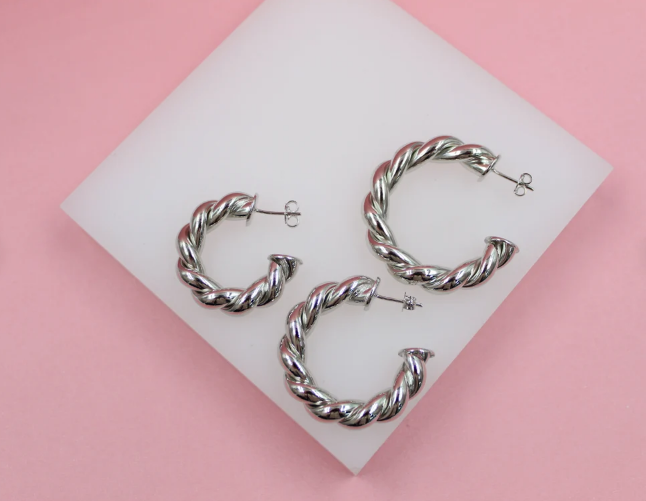 Three silver twisted hoop earrings rest on a white display stand against a pink background.