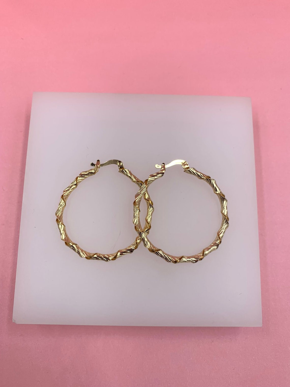 A pair of textured gold hoop earrings rests on a white display tile against a vibrant pink background.