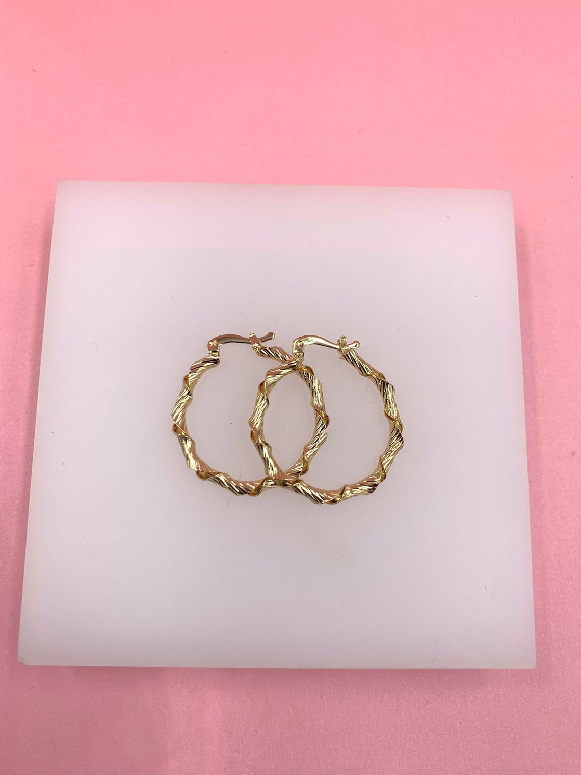 A pair of gold twisted hoop earrings rests on a white display block against a pink background.
