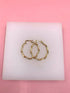 A pair of gold twisted hoop earrings rests on a white display block against a pink background.