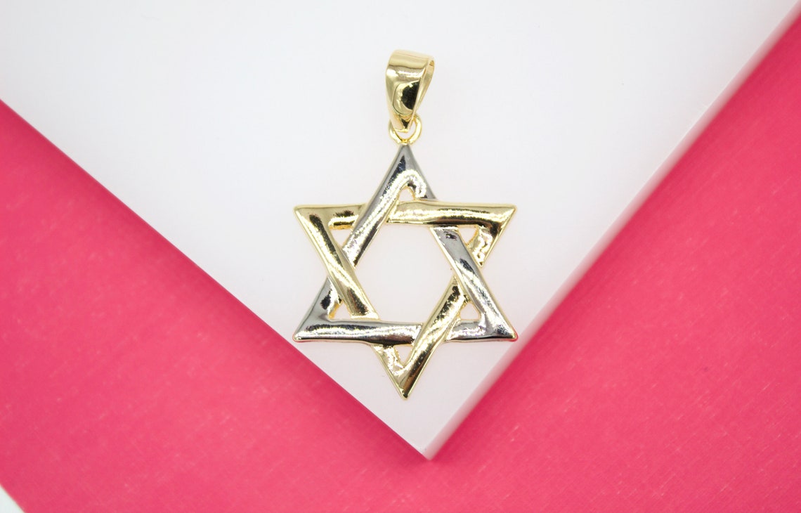 A two-tone gold and silver Star of David pendant rests on a white display surface against a bright pink background.