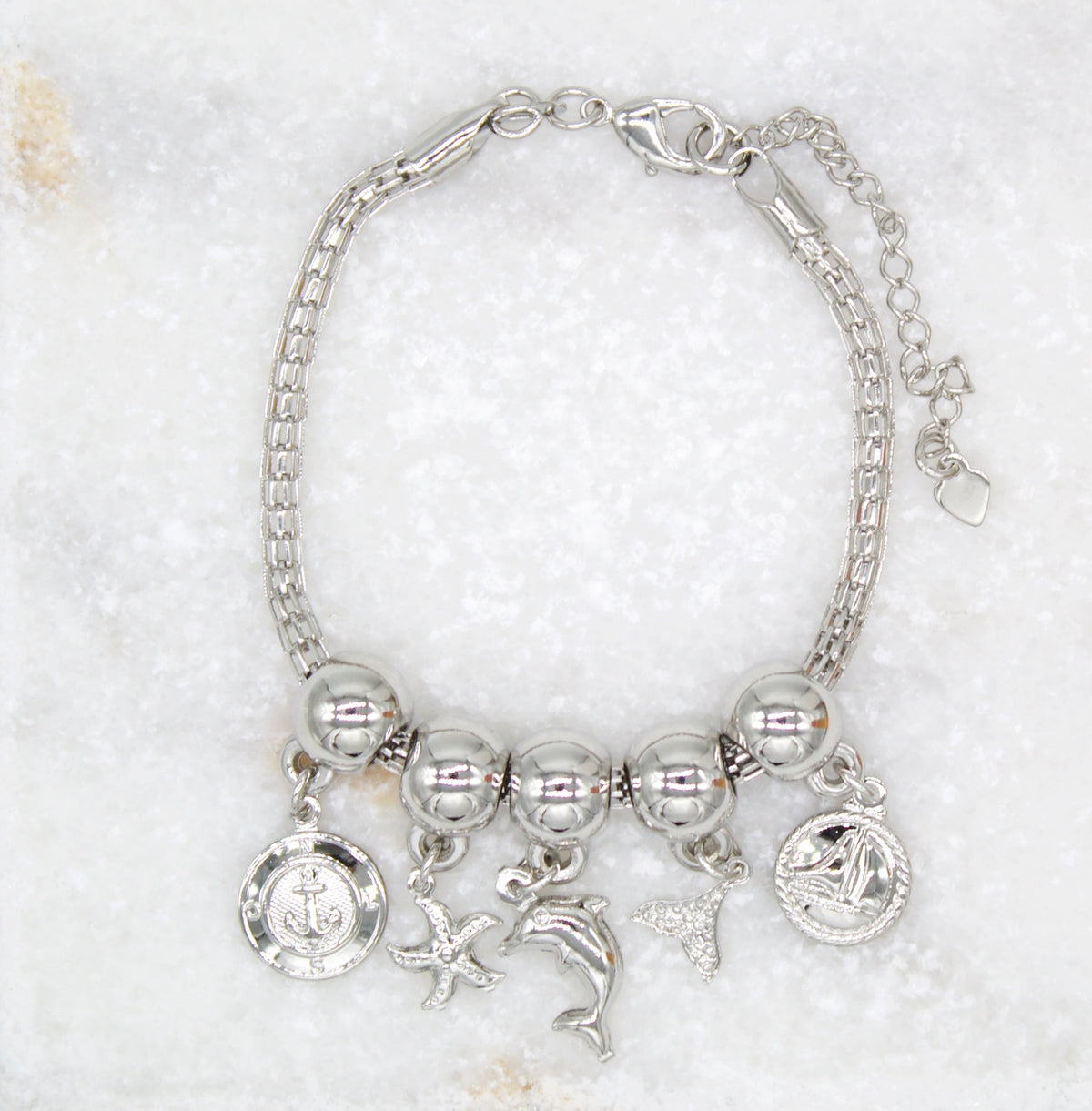 A silver nautical charm bracelet displays hanging charms of an anchor, starfish, dolphin, tail fin, and sailboat against a marble background.