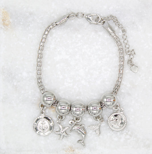 A silver nautical charm bracelet displays hanging charms of an anchor, starfish, dolphin, tail fin, and sailboat against a marble background.