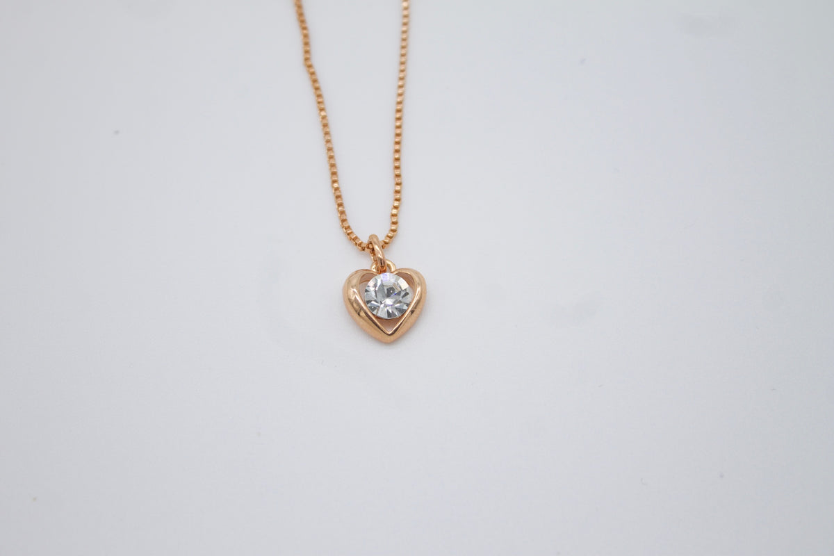 A delicate rose gold heart necklace features a brilliant crystal gemstone pendant suspended from a fine chain against a light background.