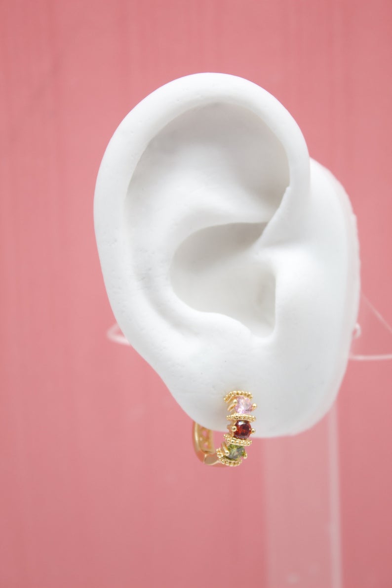 A white mannequin ear displays a gold huggie hoop earring adorned with a row of pink, red, and green gemstones against a pink background.