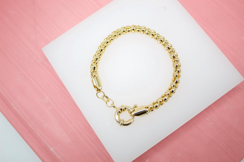 A polished gold link bracelet rests on a white display stand against a soft pink background.