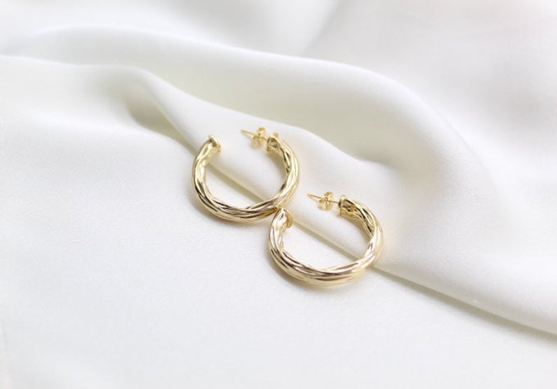 A pair of elegant gold twisted hoop earrings rest on a soft, draped white silk fabric background.