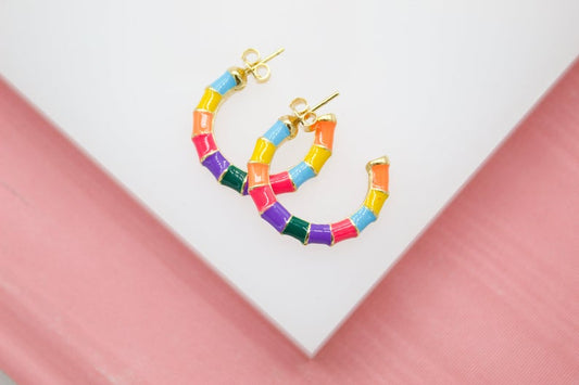 A pair of vibrant multicolored enamel gold hoop earrings rests on a white and pink surface.