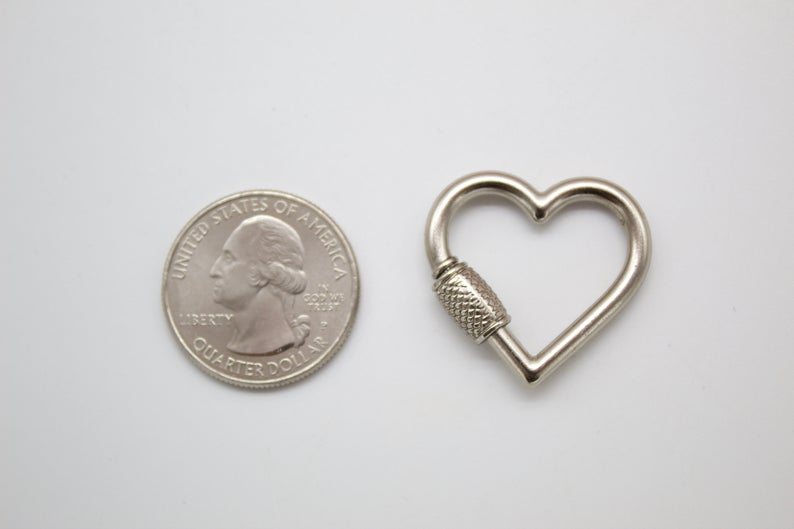 A silver heart-shaped screw-lock carabiner rests beside a US quarter for size comparison on a plain white surface.