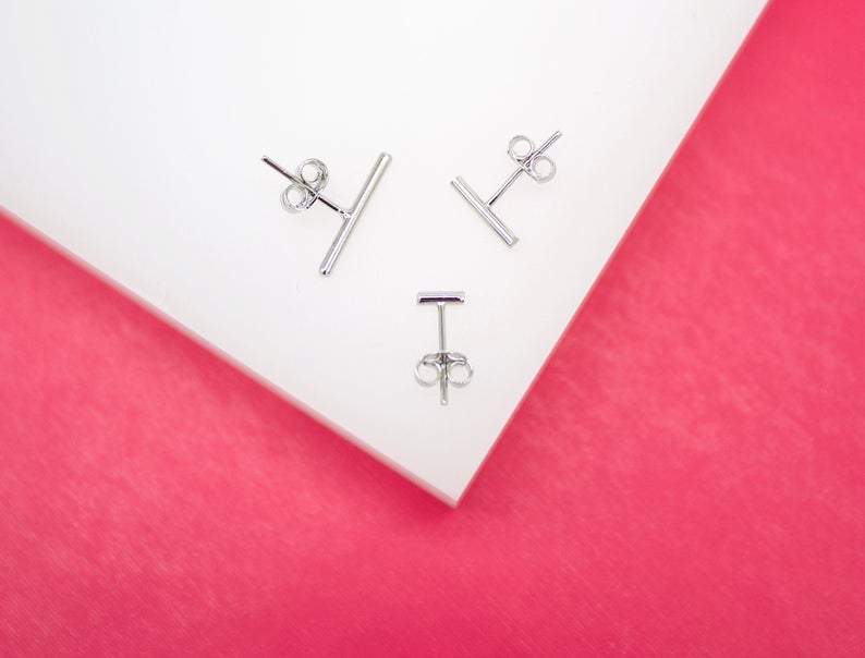 Three delicate silver bar stud earrings rest on a white surface against a vibrant pink background.