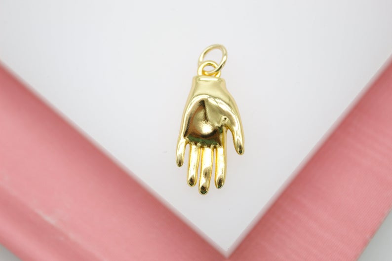 A polished gold hand charm rests on a white and pink background, showcasing its detailed palm and dangling ring.