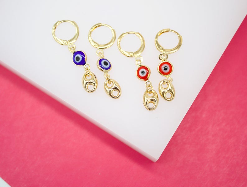 Four gold evil eye hoop earrings with dangling charms rest on a white display surface against a pink background.