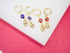 Four gold evil eye hoop earrings with dangling charms rest on a white display surface against a pink background.