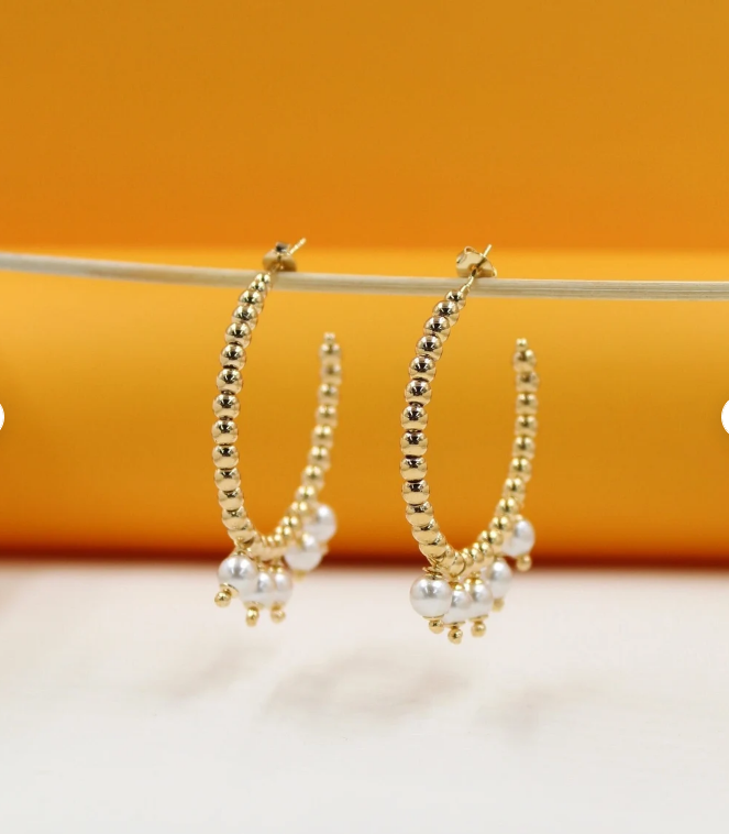 A pair of gold beaded hoop earrings featuring hanging white pearls dangles from a thin wooden stick against a warm orange and white background.