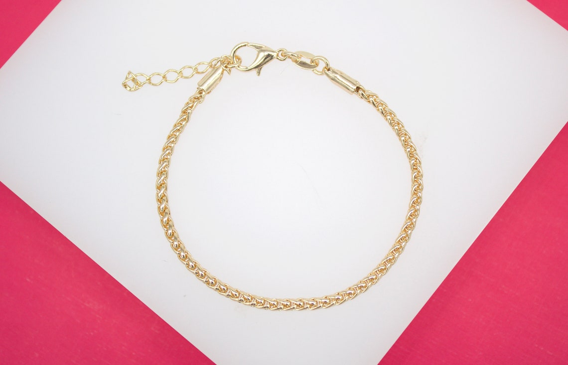 A gold wheat chain bracelet rests on a white and pink background, featuring a secure lobster clasp and an adjustable extender chain.