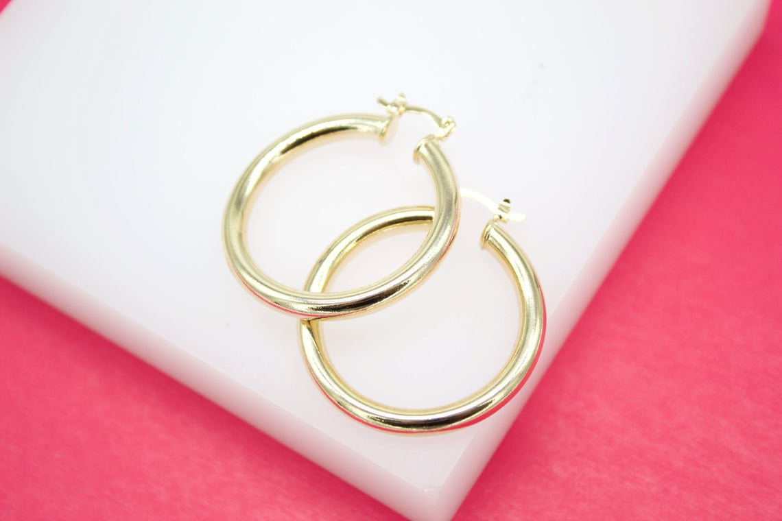 A pair of polished gold hoop earrings rests gracefully on a white display stand against a vibrant pink backdrop.
