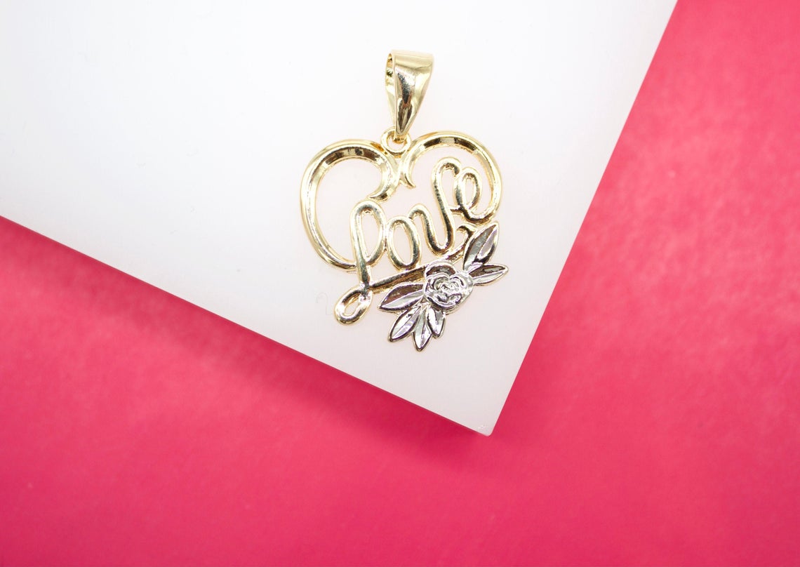 A gold heart-shaped pendant featuring the word love and a silver flower rests on a white and pink background.