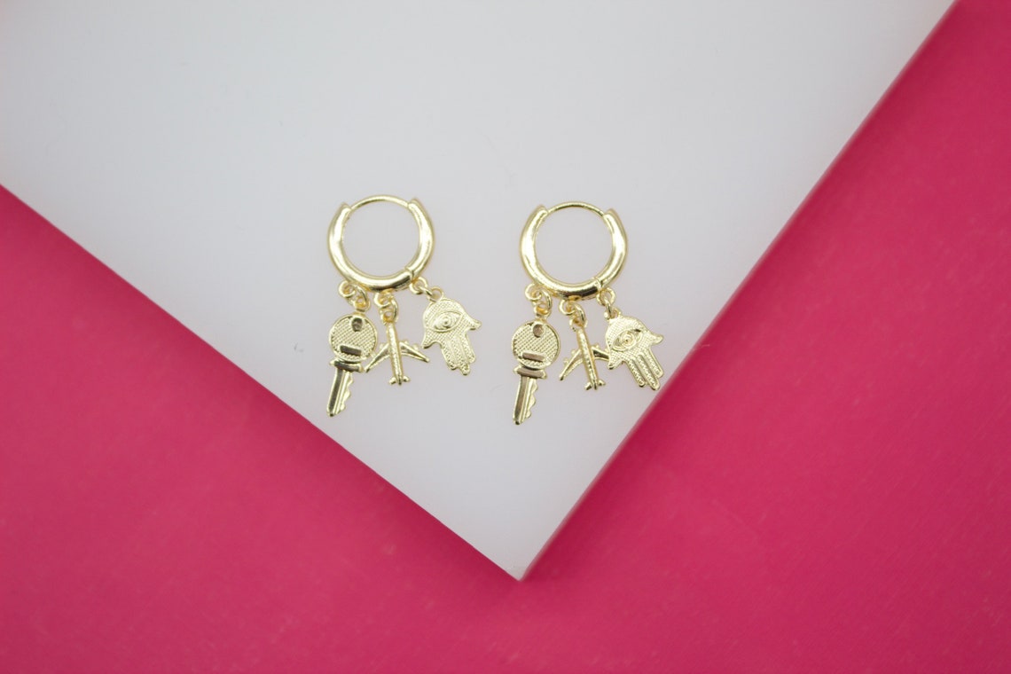 A pair of gold hoop earrings features hanging charms shaped like a key, an airplane, and a hamsa hand against a white and pink backdrop.