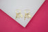 A pair of gold hoop earrings features hanging charms shaped like a key, an airplane, and a hamsa hand against a white and pink backdrop.