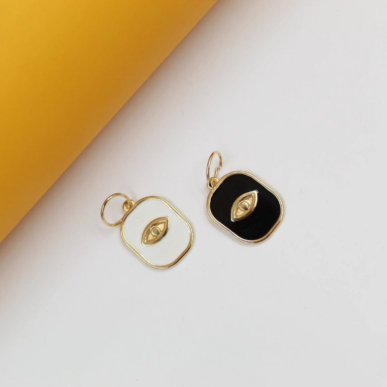 Two gold rectangular pendants with black and white enamel evil eye designs rest on a white surface next to a yellow background.