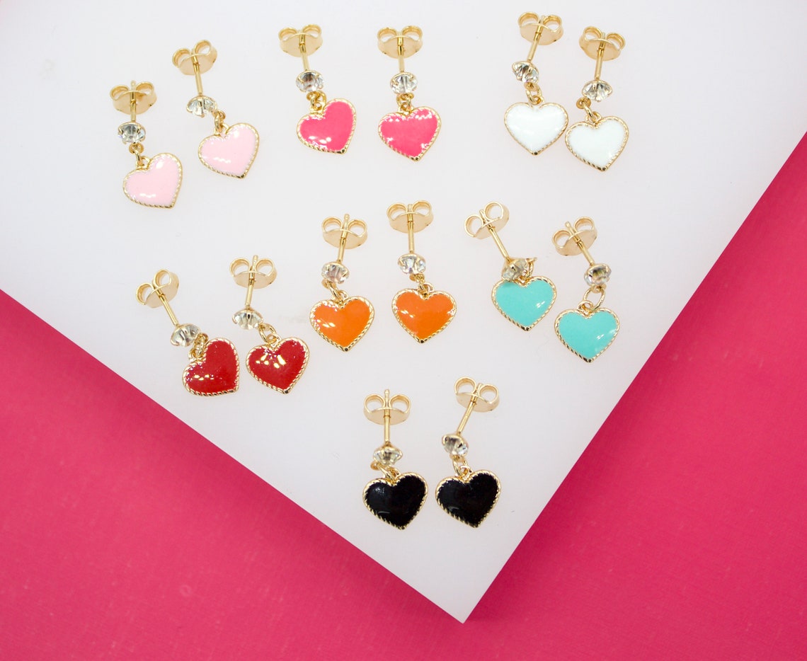 A flat lay showcases seven pairs of colorful enamel heart-shaped stud earrings with gold hardware arranged neatly on a white and pink background.