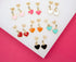 A flat lay showcases seven pairs of colorful enamel heart-shaped stud earrings with gold hardware arranged neatly on a white and pink background.