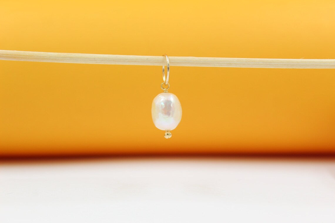 A delicate freshwater pearl charm hangs from a thin gold hoop earring against a warm orange and white backdrop.